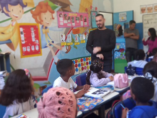 Foto de crianças sentadas em carteiras escolares em uma sala de aula. Há um homem adulto em pé